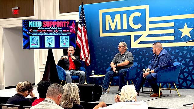 Three panelists seated on a conference stage discuss veteran suicide before an audience, with an American flag and a screen showing Veterans Crisis Line contacts behind them